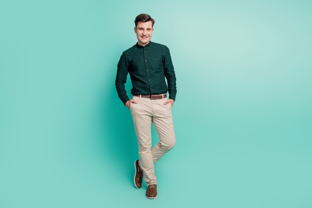 Smiling man in a dark green shirt and beige pants with brown shoes posing casually against a teal background with hands in pockets.