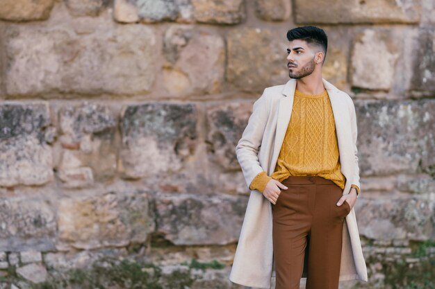 Man in a stylish beige coat, mustard sweater, and brown trousers posing outdoors against a rustic stone wall background.